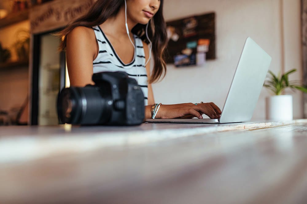 Woman blogger using laptop at home wearing earphones. Woman sitting with DSLR camera on the table working on her laptop computer.