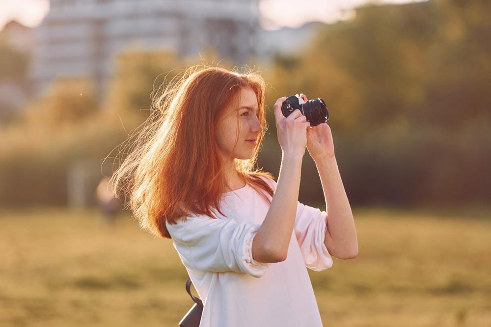 Photographer is with camera. Young girl is on the field at sunny daytime having nice weekend.